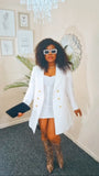 Woman in an all white coat and white inner wear standing in a room with a chandelier and framed pictures on the wall.