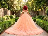 A woman viewed from behind wearing a voluminous peach tulle ball gown with an off-the-shoulder neckline and a long train, standing on a stone path in a garden with hedges and trees.