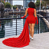 A woman stands on a waterfront promenade wearing a sparkling red sequined mini dress with a long train, matching red gloves, and white heels, photographed from behind.