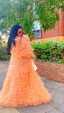 Woman in a peach ruffled dress standing outdoors with greenery and a building in the background