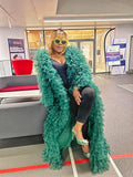 Woman wearing a green ruffled coat sitting on a chair in an indoor setting.