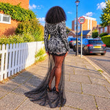 A woman stands on a sunny sidewalk in a residential street, photographed from behind, wearing a black and white patterned long-sleeve mini dress with sheer black tights and a long embellished train.