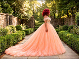 A woman stands outdoors on a stone path wearing a voluminous peach ball skirt and a long train, photographed from the back in a garden setting.