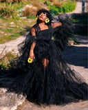 A woman wearing a dramatic black tulle ball gown with sheer sleeves and voluminous layered ruffles, standing outdoors on a stone path with greenery in the background. She is holding yellow flower petals and wearing large yellow flower earrings.
