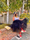 Woman in a black tulle skirt and red sandals sitting on a brick wall outdoors.